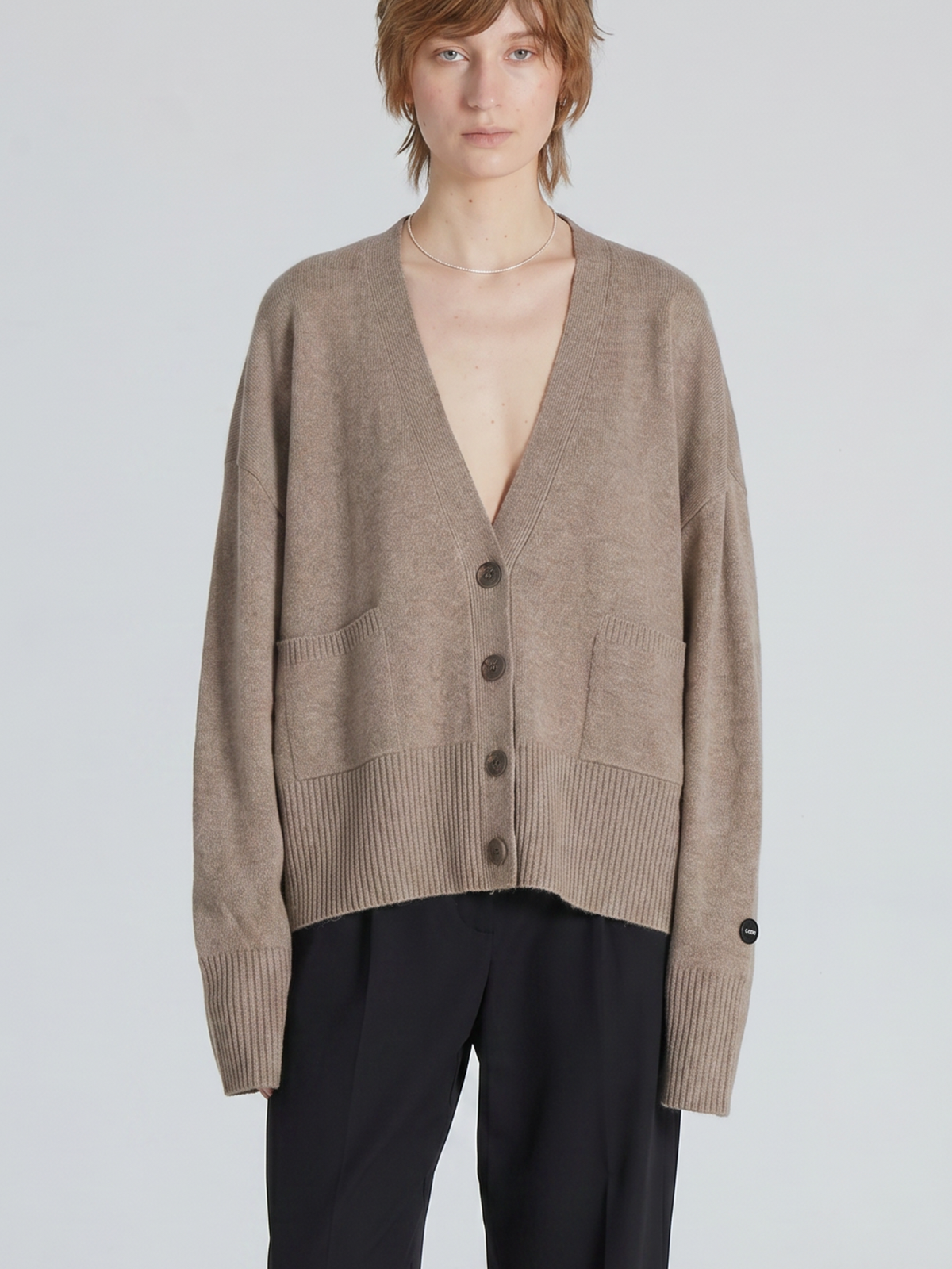 A model wearing a taupe brown knit cardigan with a deep V-neck, front buttons, and front patch pockets, paired with black trousers, against a light gray studio background.