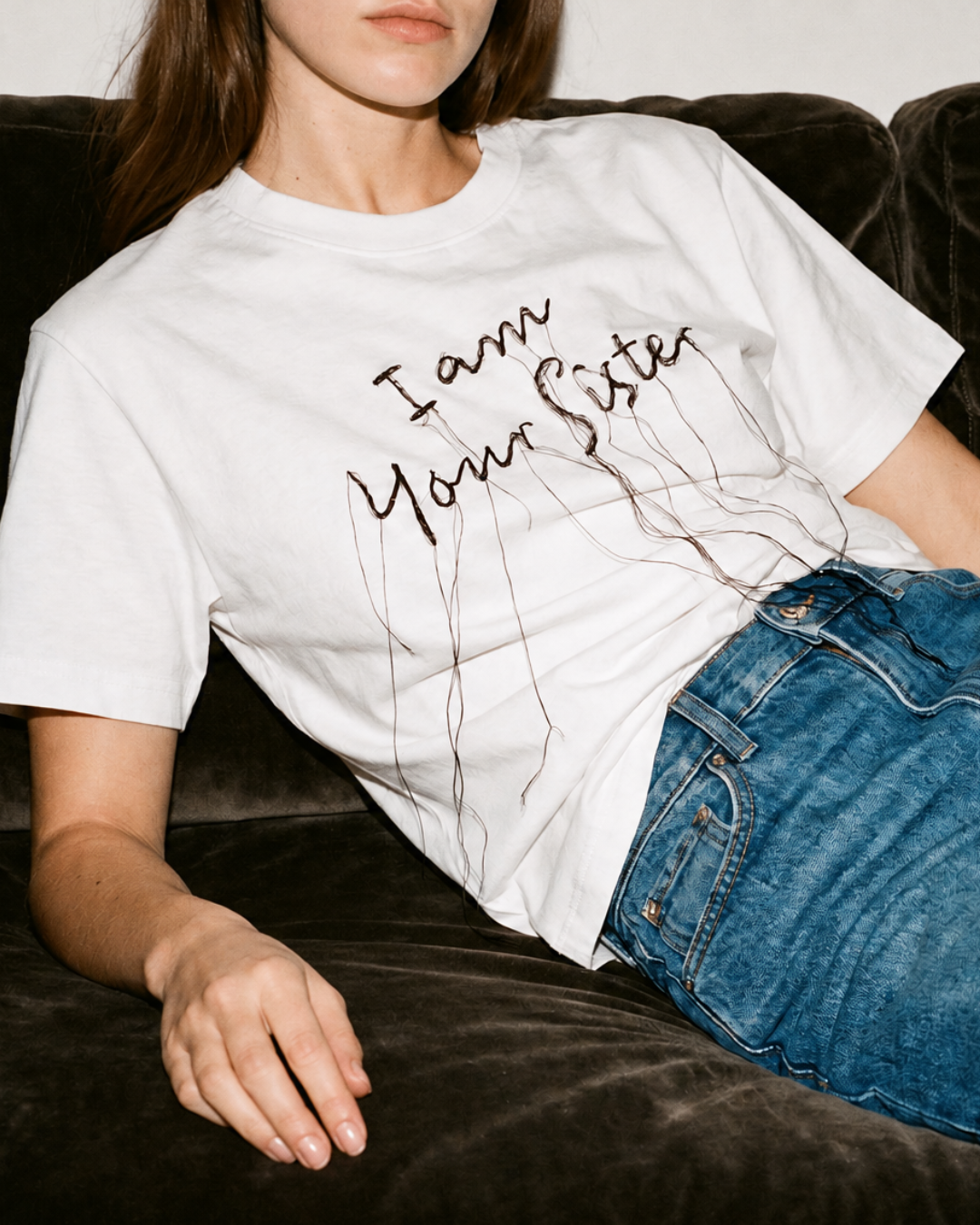 A woman wearing a white graphic t-shirt with the text 'I am your sister' in a dark script, styled with blue jeans while reclining on a dark brown velvet couch.
