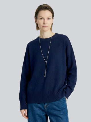 A woman is wearing a navy cashmere sweater and blue jeans on a light gray background