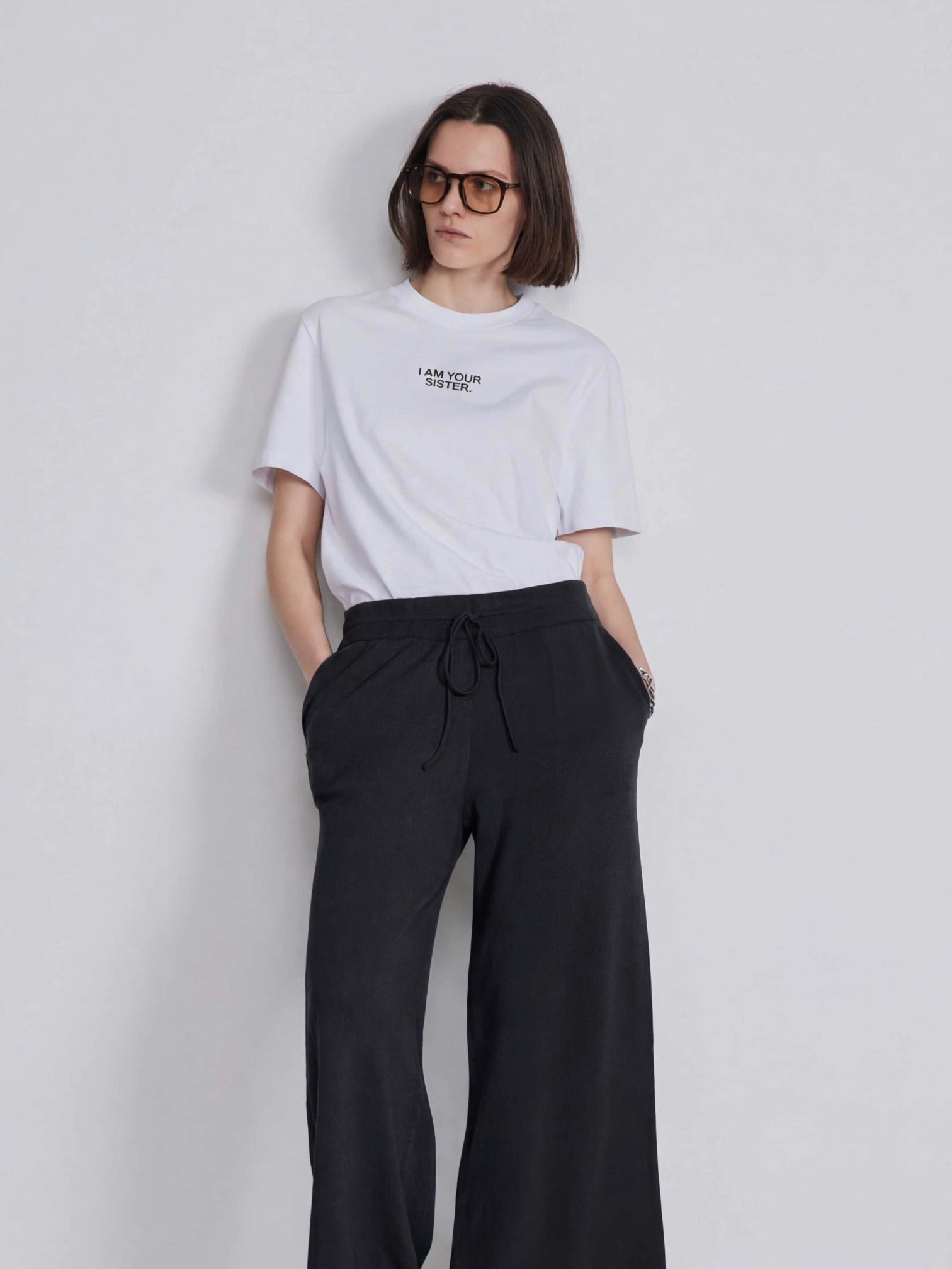 Woman wearing a white Sister t-shirt and black pants against a plain background
